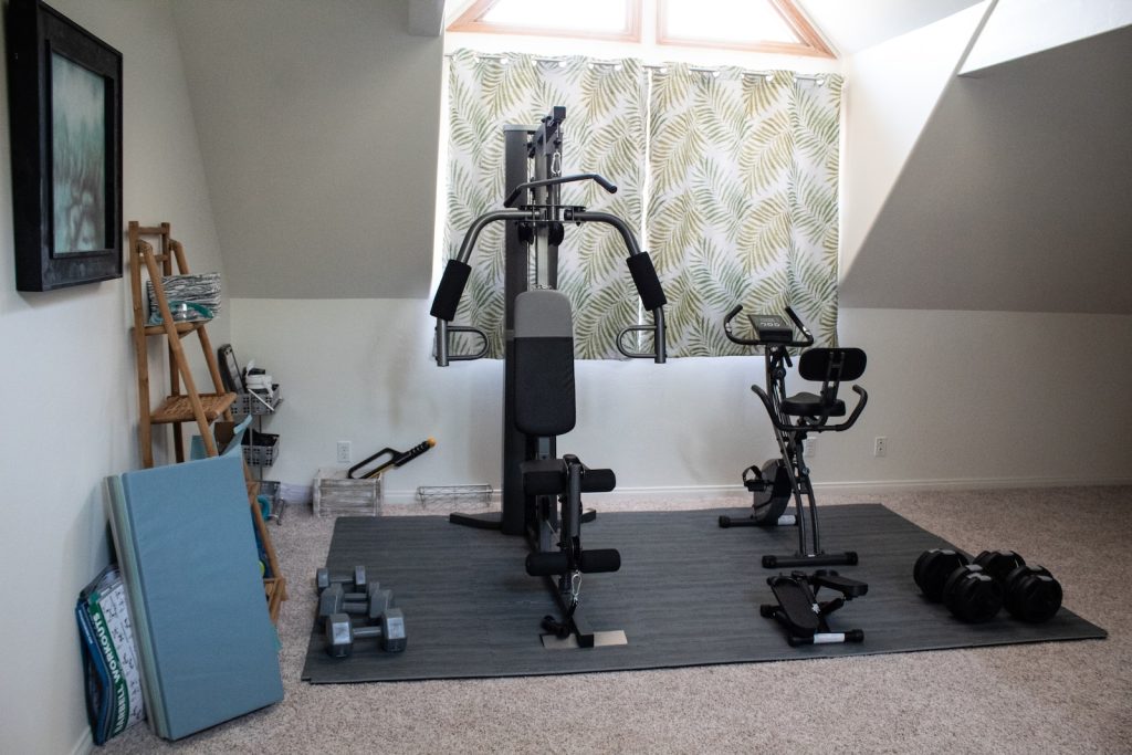 Home gym with exercise bike, weight machine, dumbbells, and yoga mats in a well-lit attic space with decorated curtains