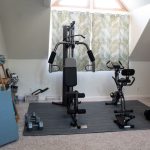 Home gym with exercise bike, weight machine, dumbbells, and yoga mats in a well-lit attic space with decorated curtains