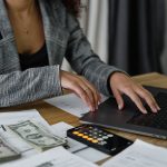 Person in blazer working on laptop with calculator and dollar bills on desk