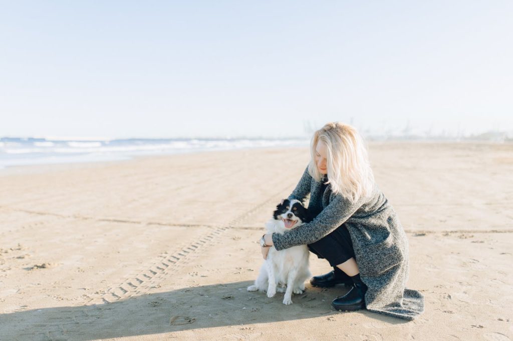Woman with blonde hair kneeling on a sandy beach, hugging a small black and white dog near the shoreline