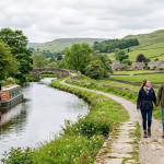 A couple walks along a scenic canal path in the Yorkshire Dales countryside with a narrowboat and stone bridge nearby