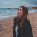 Woman in black jacket enjoying a coastal view on a sandy beach with industrial plant and cliffs in the background