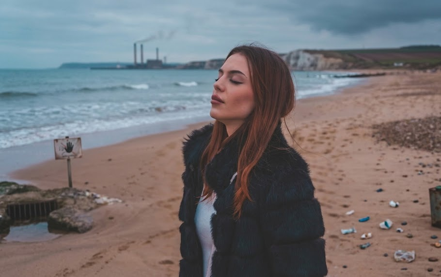 Woman in black jacket enjoying a coastal view on a sandy beach with industrial plant and cliffs in the background